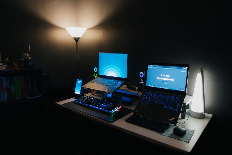 A sleek gaming desk with two laptops, RGB keyboard, and ambient lighting
